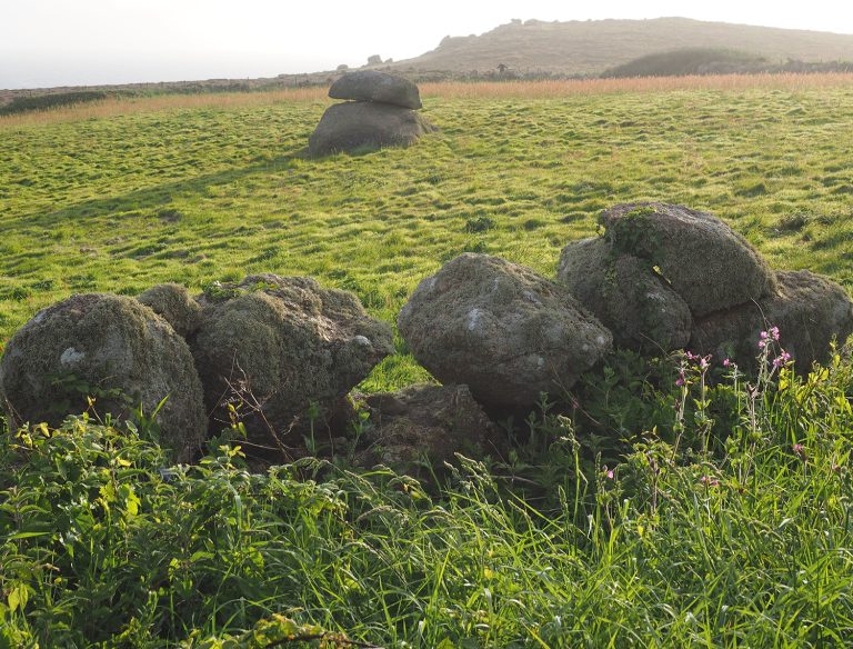 A weathered stone hedge covered in lichen and moss stands in a green coastal field, with a balanced granite boulder formation visible in the misty distance